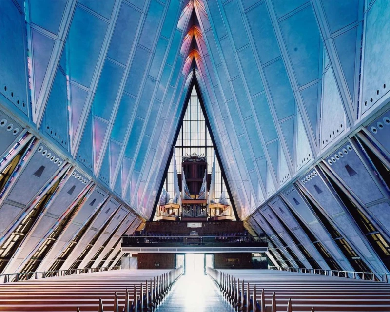 Interior view of a modern chapel with rows of pews leading to a large pipe organ in the background, featuring geometric architectural details and blue lighting.