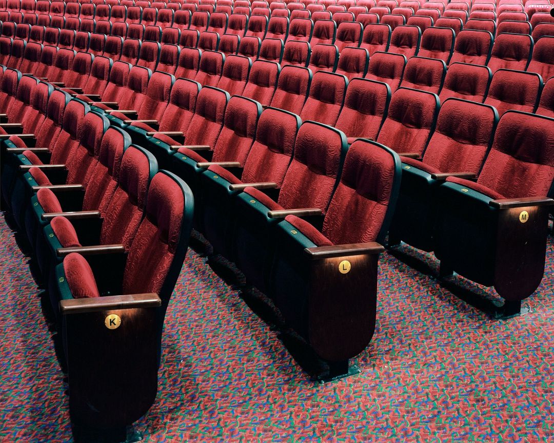 Rows of empty red cushioned seats in a theater or auditorium, arranged in neat lines with colorful patterned flooring.