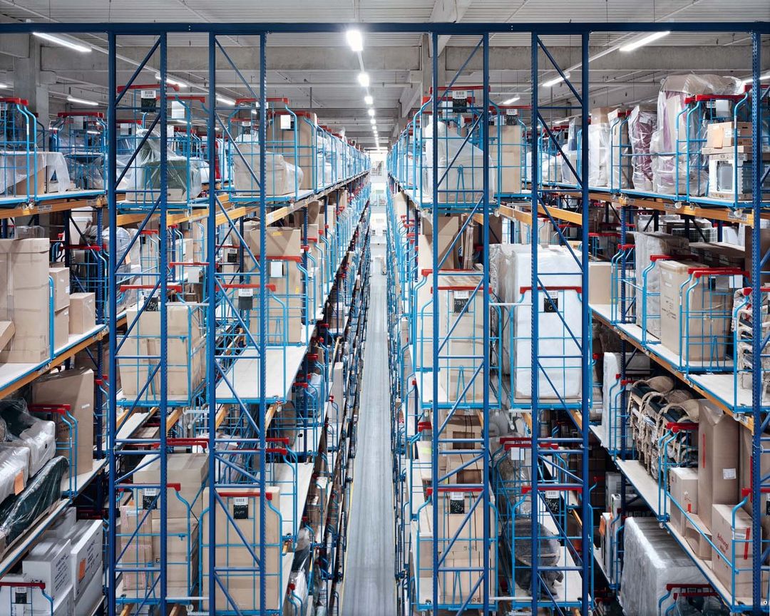 Warehouse interior with rows of high storage racks holding boxes and goods, lit by overhead lighting.