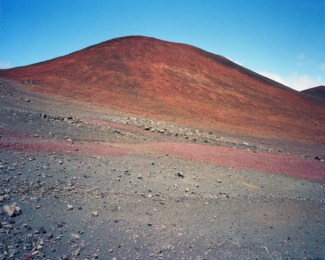 Red sand and rocks landscape with a large hill under a clear blue sky, creating a textured and colorful outdoor environment.