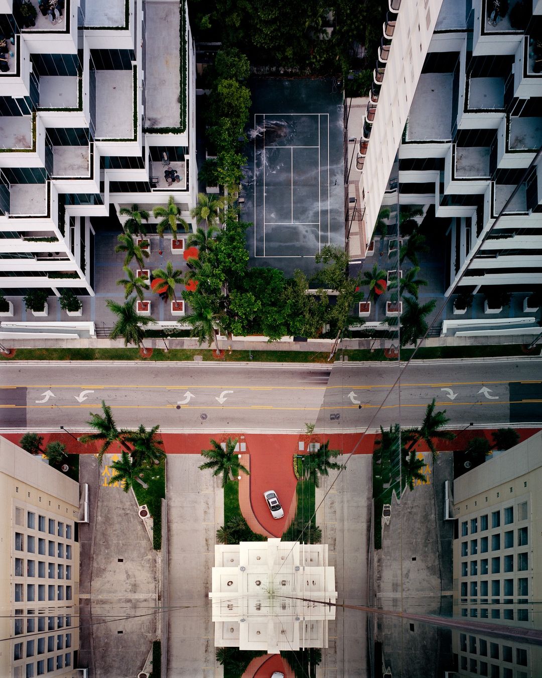 Aerial view of a modern city featuring high-rise buildings, palm trees, a tennis court, and a car parked on a red walkway.