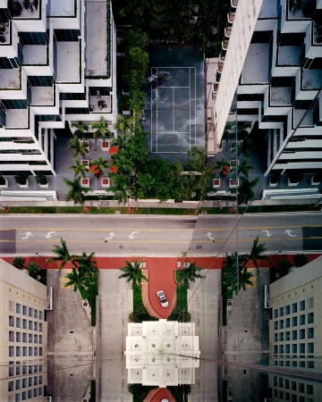 Aerial view of a modern city featuring high-rise buildings, palm trees, a tennis court, and a car parked on a red walkway.