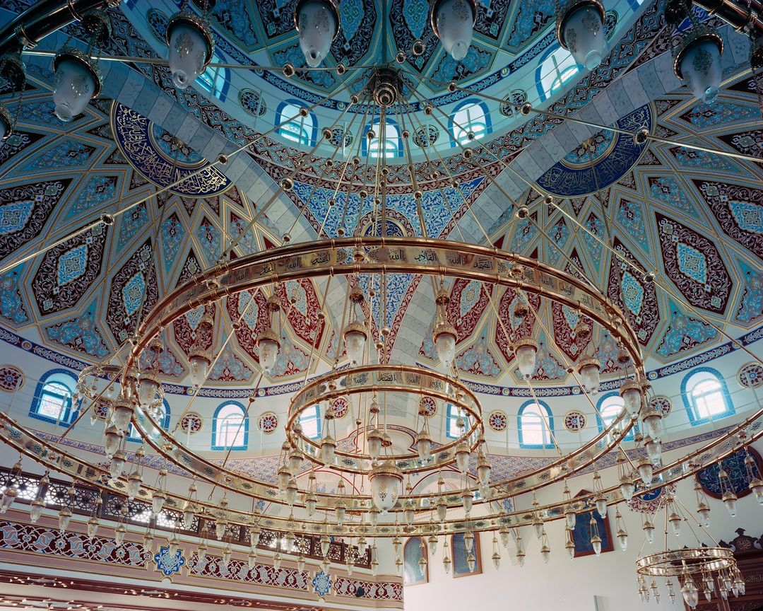 Intricate architectural dome featuring detailed geometric patterns, ornamented with chandeliers, in an interior with blue and gold colors.