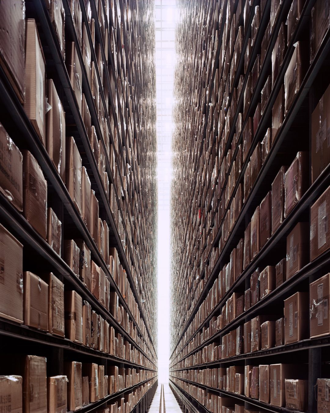 A warehouse aisle shows long rows of shelves filled with stacked boxes receding into the distance, lit by bright overhead lighting.