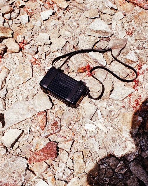 Overhead shot of a black vintage camera bag with a strap on a beige rocky surface with specks of red and brown. 