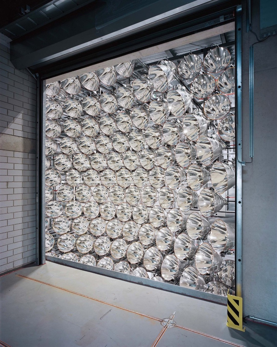 View of a warehouse interior through a partially open shutter door, with a grid of mirrored reflectors filling the space.