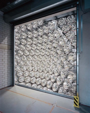 View of a warehouse interior through a partially open shutter door, with a grid of mirrored reflectors filling the space.