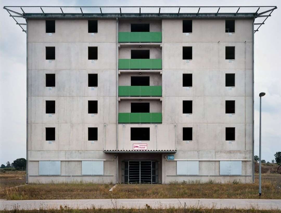 Exterior view of a concrete building with multiple window openings and a green balcony structure on a partly cloudy day.