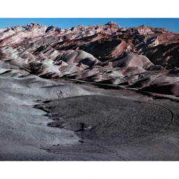 Aerial or high-angle view of barren desert terrain with undulating grey and reddish mountain ridges under a clear blue sky.
