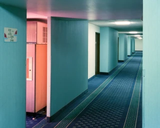 A long hotel corridor with teal walls and blue patterned carpet stretching into the distance under overhead lights.