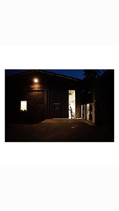 Exterior shot of a large dark warehouse or industrial building at night, illuminated by a single light source with a person visible in the doorway.