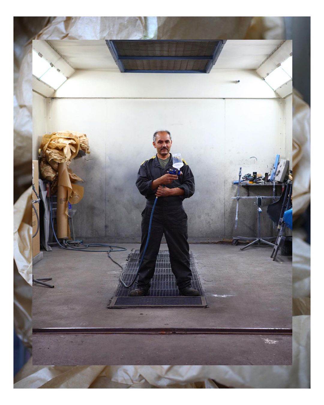 An adult male stands confidently in an indoor auto repair shop, holding a spray gun, with a focus on equipment and industrial setting.