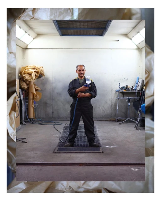 An adult male stands confidently in an indoor auto repair shop, holding a spray gun, with a focus on equipment and industrial setting.