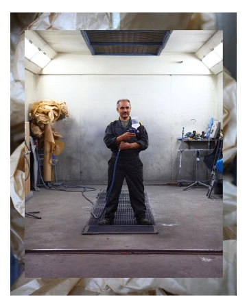 An adult male stands confidently in an indoor auto repair shop, holding a spray gun, with a focus on equipment and industrial setting.
