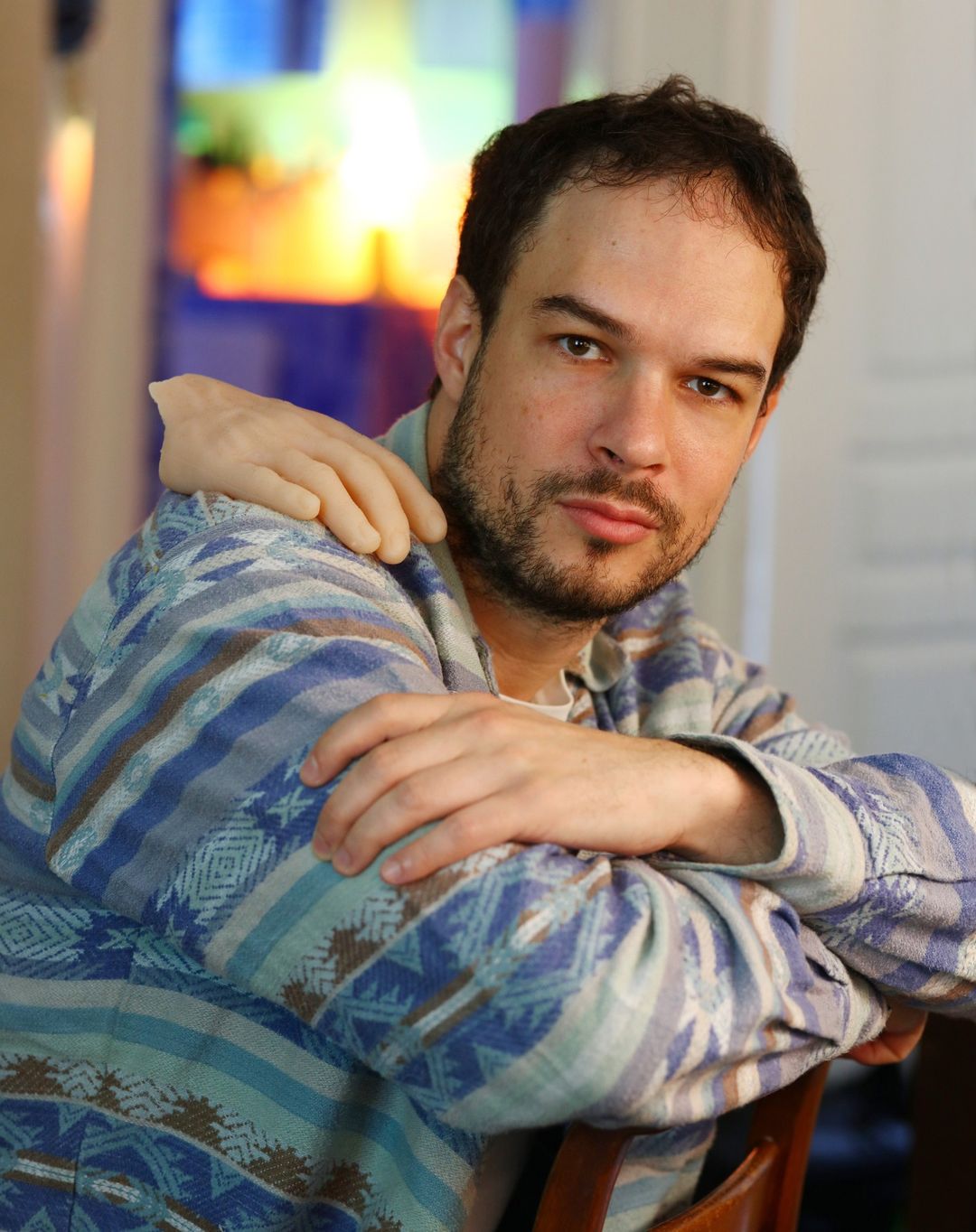 A man with dark hair is posed indoors wearing a patterned shirt, with a realistic prosthetic hand resting on his shoulder. Portrait