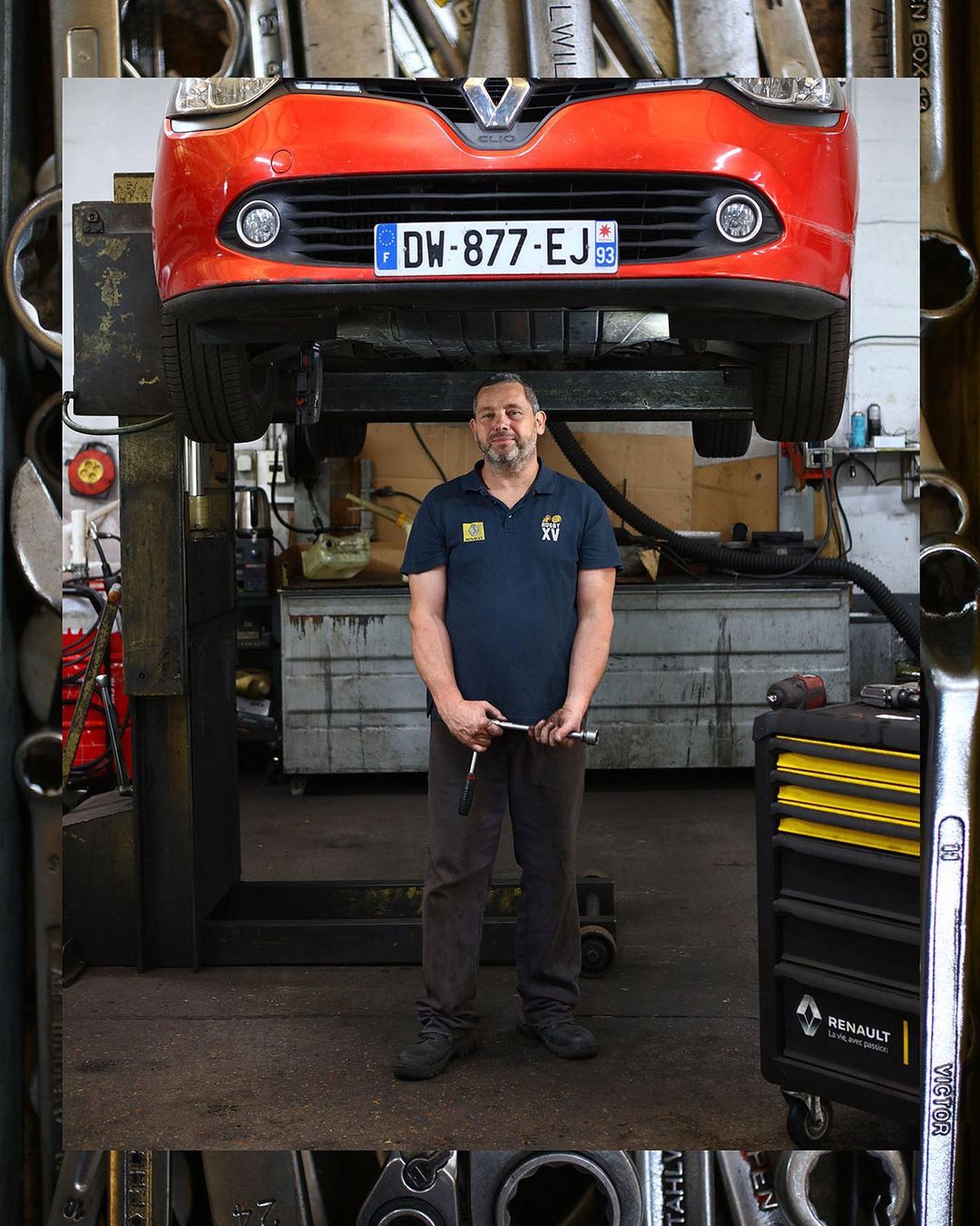 Auto mechanic stands in a garage with a raised red car, holding tools, surrounded by tools and equipment, capturing an automotive setting.