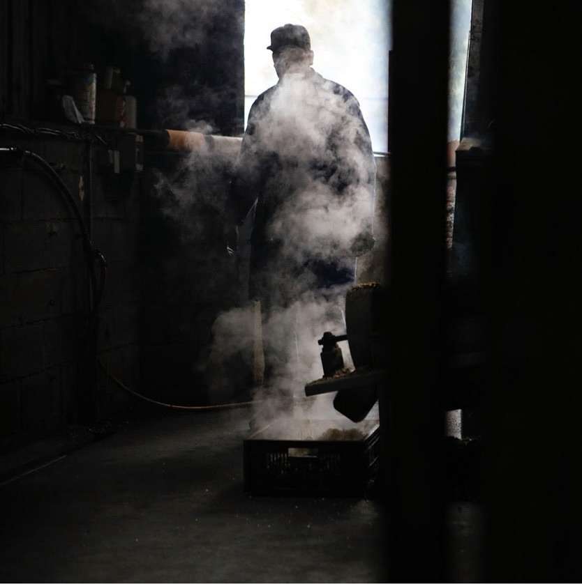 Man in hat and coat stands in industrial setting with steam, dark room, and equipment. Moody, gritty image with strong contrast.