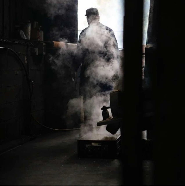 Man in hat and coat stands in industrial setting with steam, dark room, and equipment. Moody, gritty image with strong contrast.