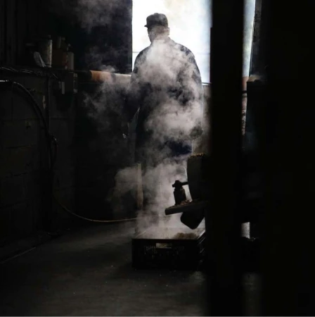 Man in hat and coat stands in industrial setting with steam, dark room, and equipment. Moody, gritty image with strong contrast.