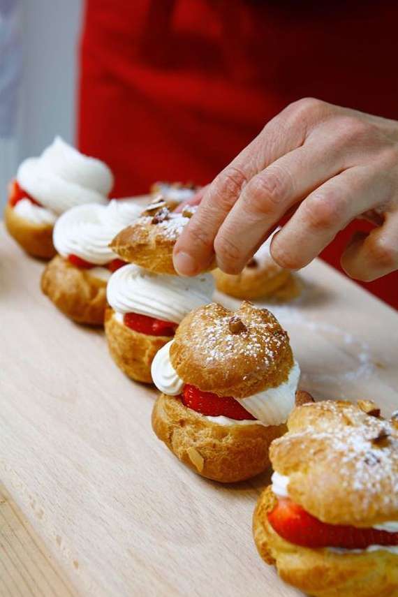 A hand carefully places the top of a cream puff on top of whipped cream and a strawberry slice. Confectioneries are on a wooden board.