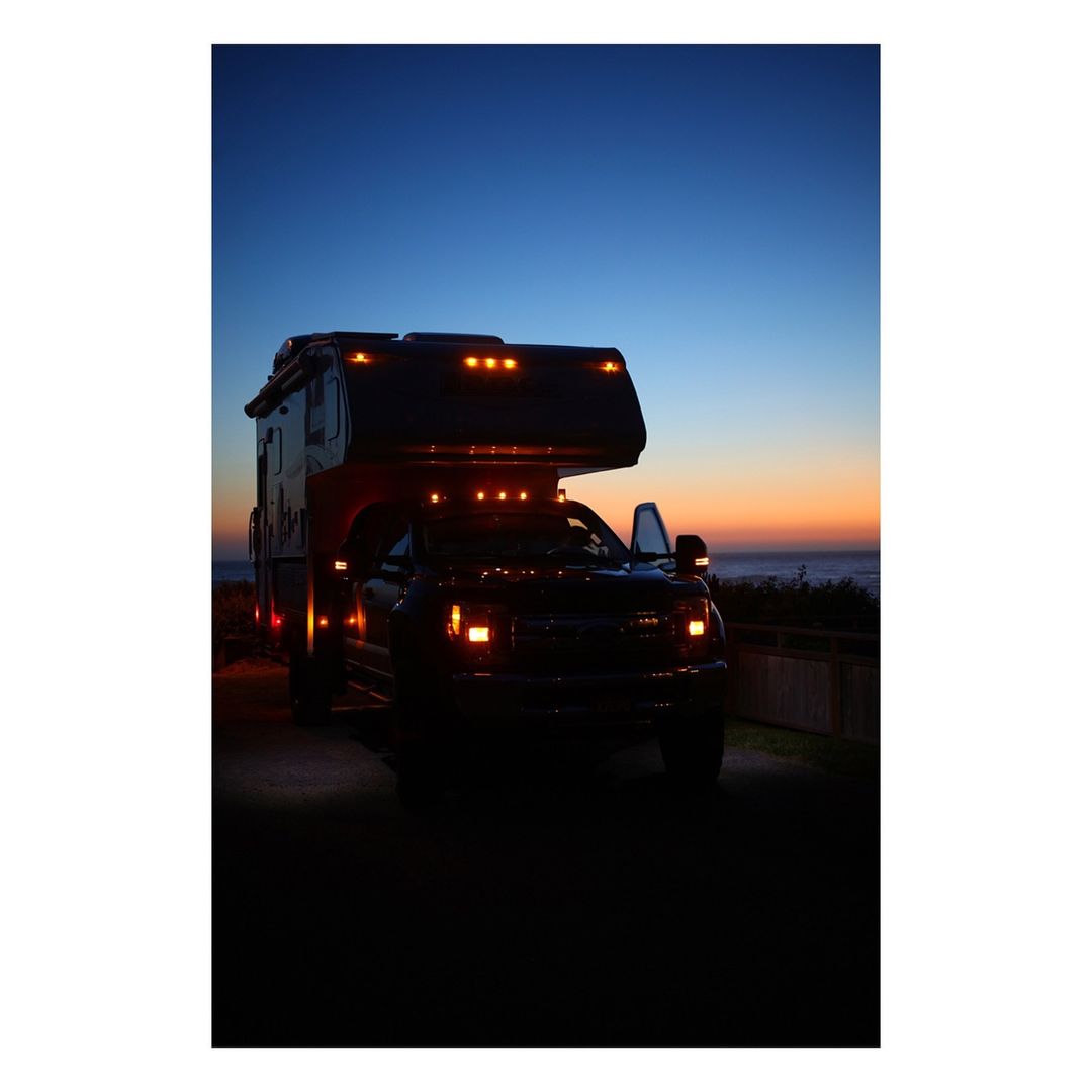 A dark pickup truck camper is parked by the beach, lit by its headlights and marker lights, set against a vibrant, colorful sunset sky.