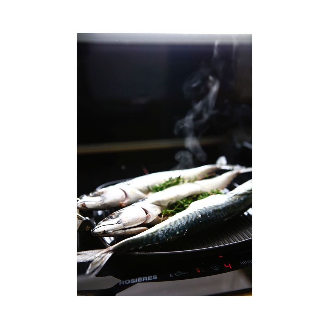 Three fresh mackerels are arranged on a black stovetop with sprigs of green herbs and rising steam, set against a dark background.