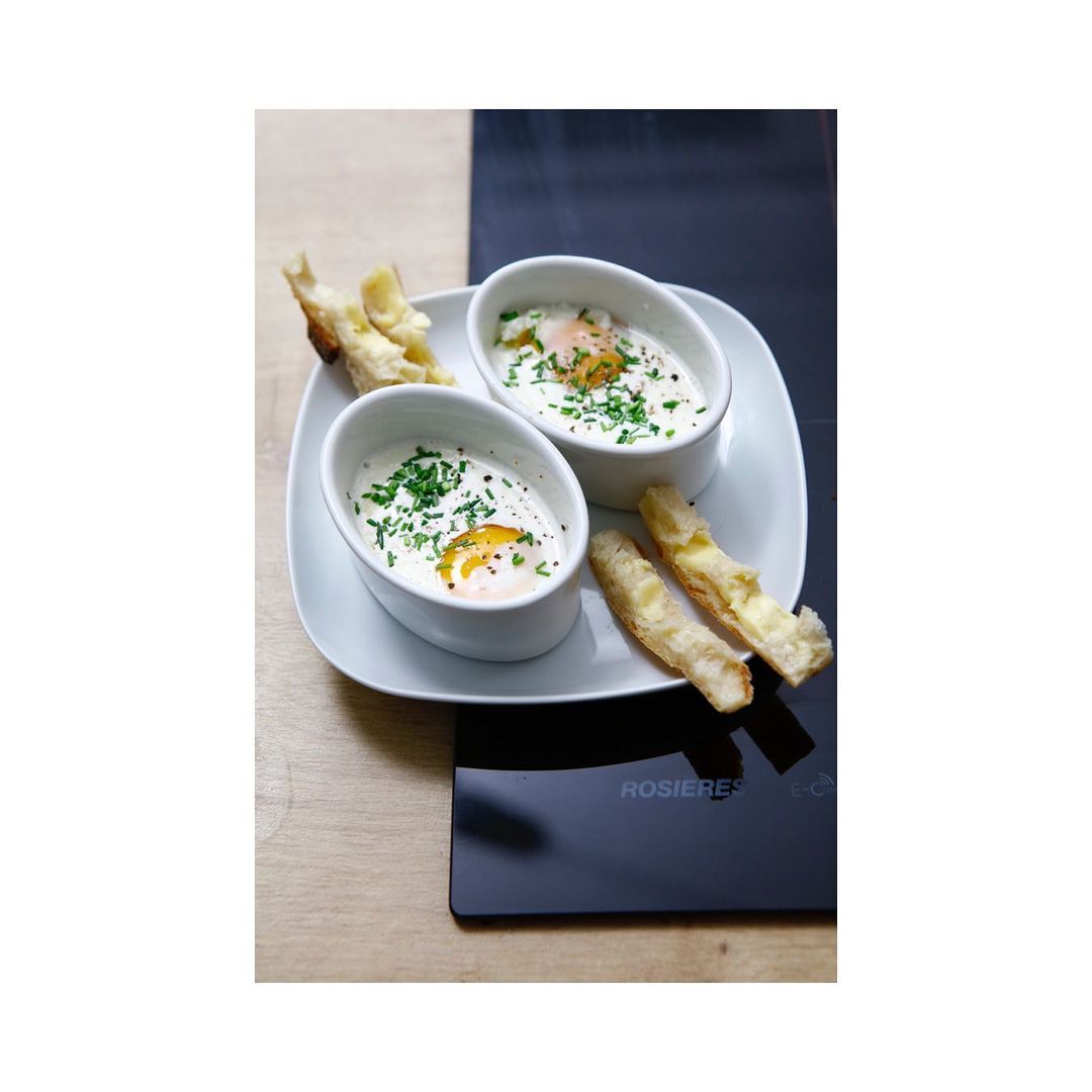 Overhead view of two white ramekins with baked eggs and herbs, served with toasted bread on a square plate, with a black surface nearby.