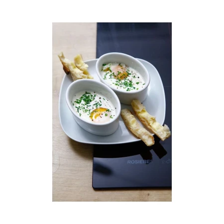 Overhead view of two white ramekins with baked eggs and herbs, served with toasted bread on a square plate, with a black surface nearby.