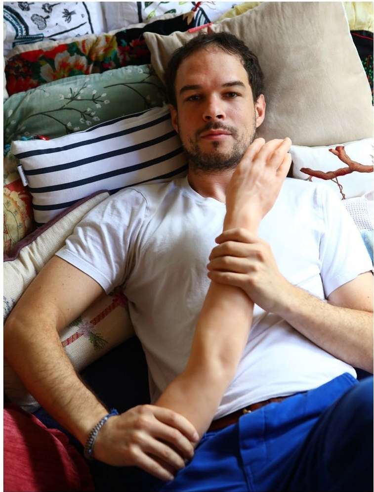 Adult male rests on an assortment of pillows in an indoor setting, wearing a white t-shirt and blue pants, holding an arm.