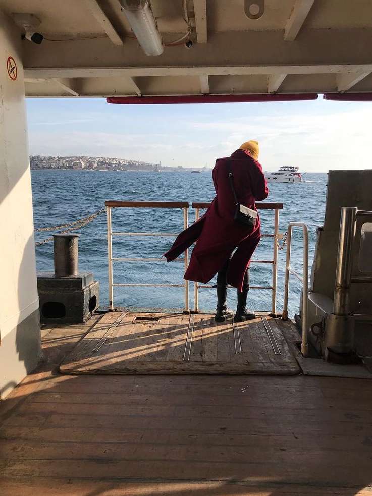 Woman in red coat stands on a ferry, looking out at the water with distant city skyline, providing a travel and lifestyle feel.
