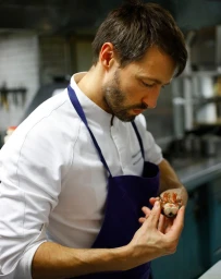 A chef in a white chef jacket and blue apron examining a small dish in a professional kitchen
