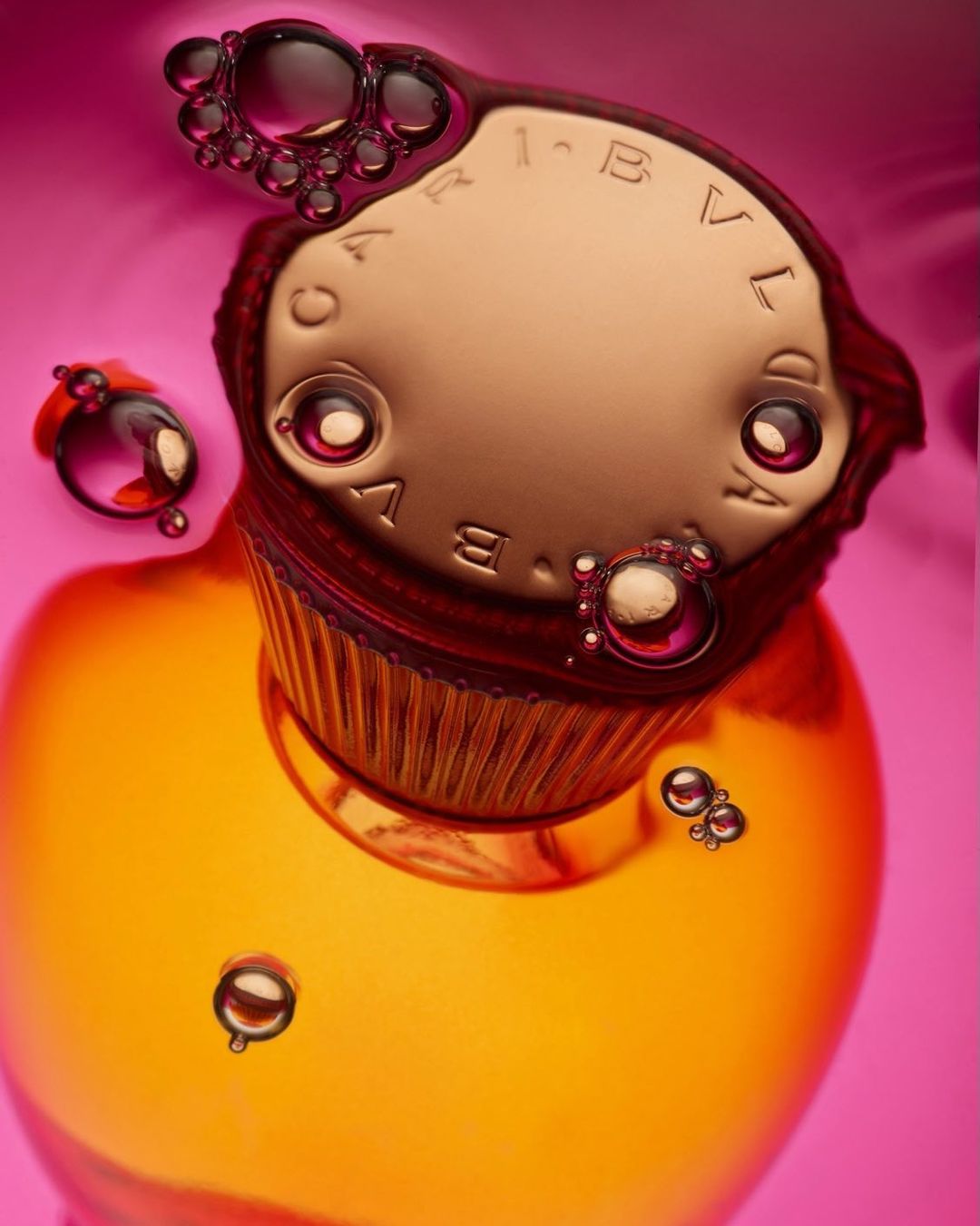 Close-up product shot of a gold perfume bottle partially submerged in pink liquid, covered in air bubbles. Branding text is visible.