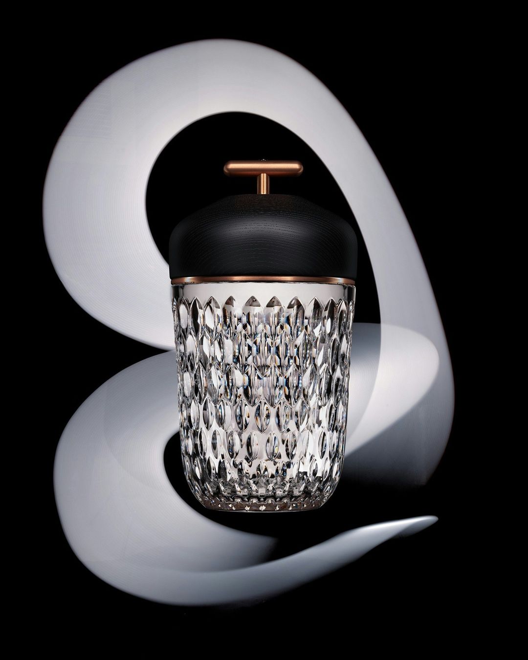 Crystal ice bucket with black leather lid and copper handle in a studio setting with white swirl and dark background.