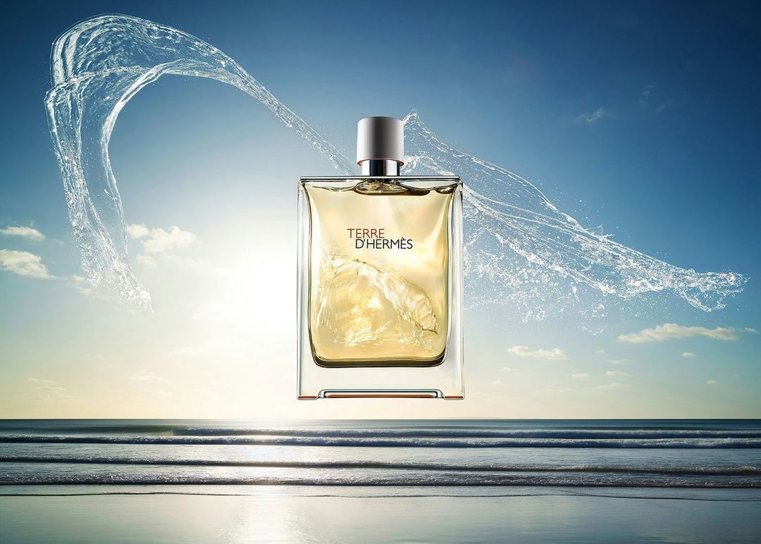 Terre d'Hermès fragrance bottle floats above a calm ocean during sunset, framed by water splashing and clear blue sky.