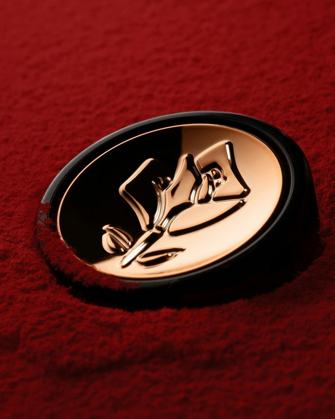 Close-up of a gold-colored Lancôme brand emblem featuring a stylized rose, placed on a textured red powder-like background.