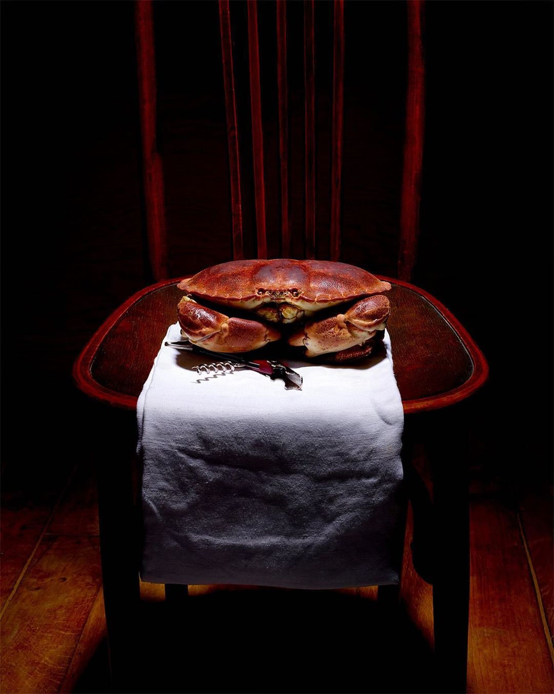 Still life of a crab served on a wooden chair with a white cloth and a wine opener, set against a dark background with red accents.