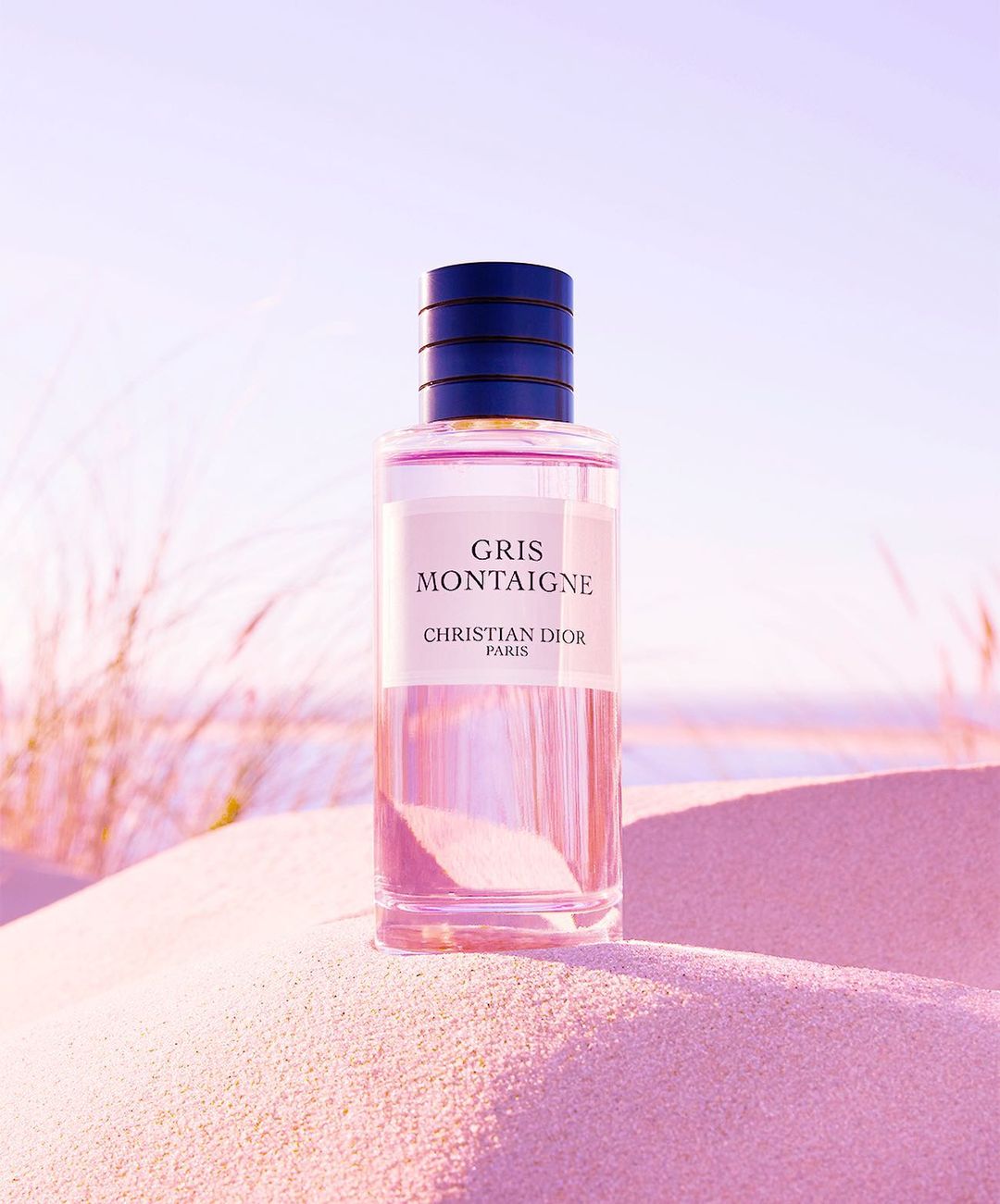 A perfume bottle of Gris Montaigne by Christian Dior sits on pink sand with blades of dune grass in the background under a light sky.