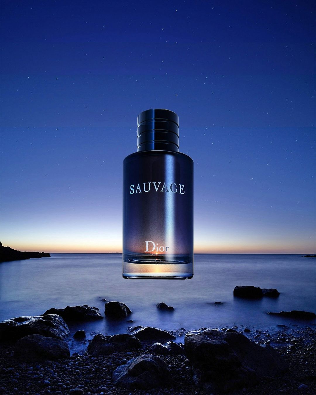 Nighttime product shot of a Dior Sauvage cologne bottle on a rocky shore with an ocean and star-filled sky background. Moody lighting.