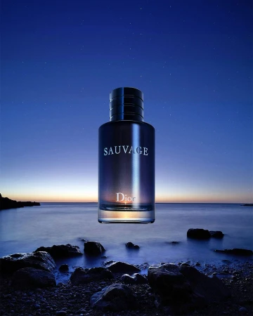 Nighttime product shot of a Dior Sauvage cologne bottle on a rocky shore with an ocean and star-filled sky background. Moody lighting.