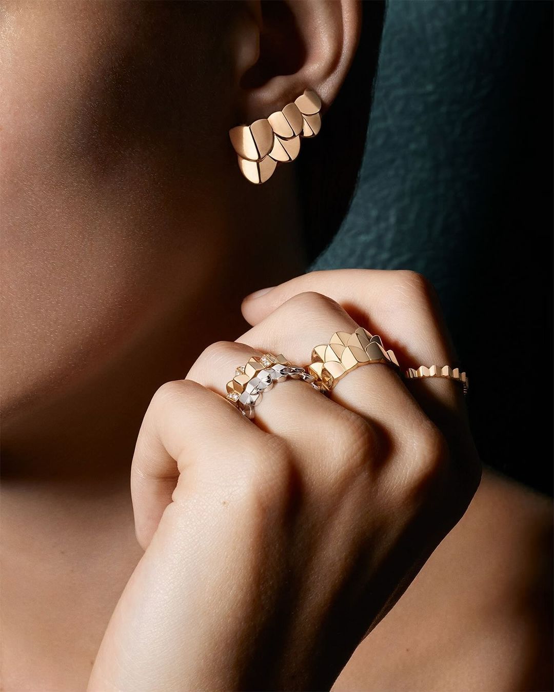 Close-up of a person's ear adorned with gold earrings, and rings with geometric shapes on their fingers, against a dark backdrop.