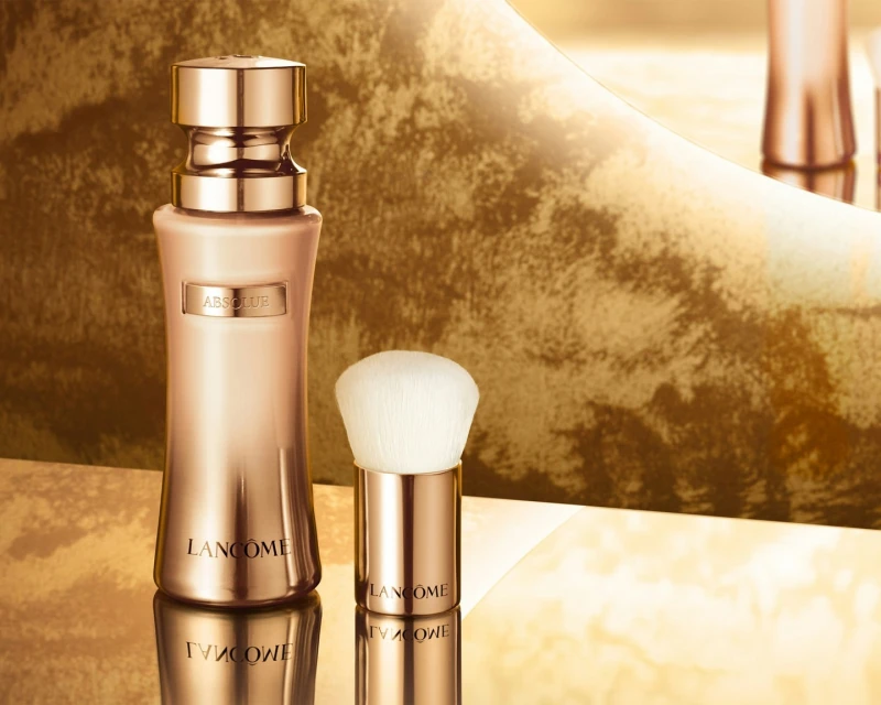 Elegant rose gold Lancôme Absolue beauty serum bottle and applicator brush, glossy reflective surface, and luxurious golden backdrop.