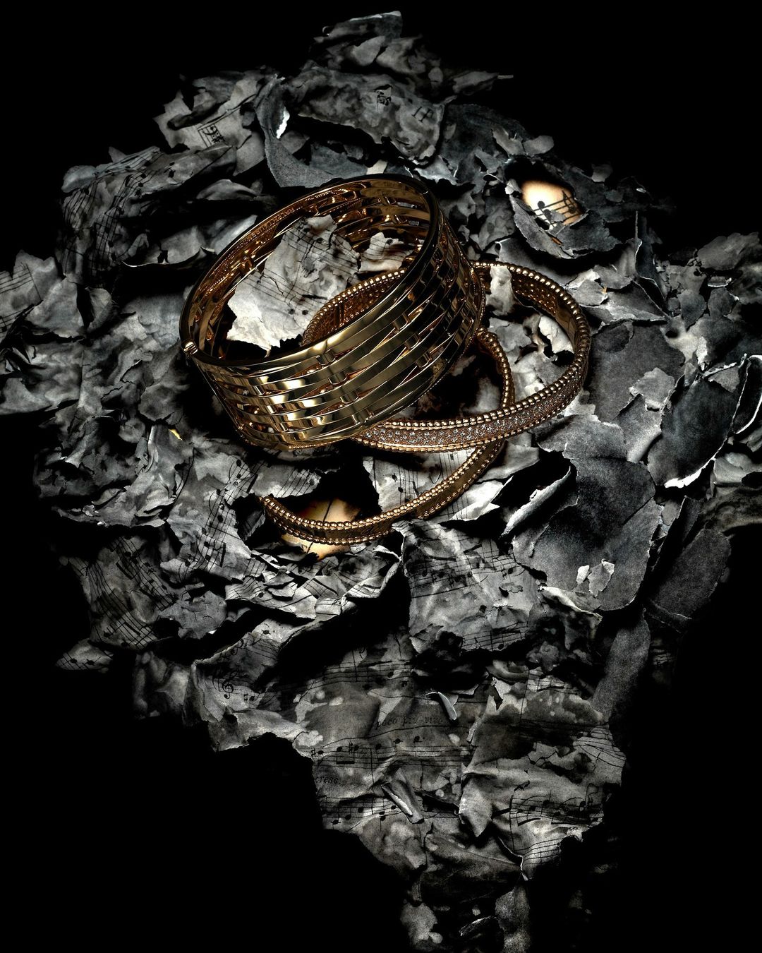 Still life of gold bracelets on a bed of burned sheet music fragments against a stark black background.