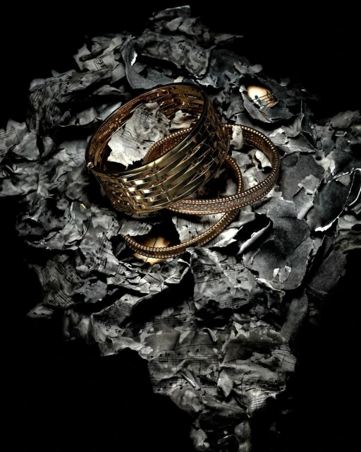 Still life of gold bracelets on a bed of burned sheet music fragments against a stark black background.