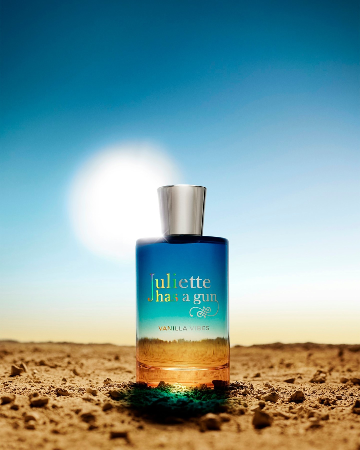 Blue and orange perfume bottle with a silver lid centered against a blue sky and desert landscape backdrop under natural lighting.
