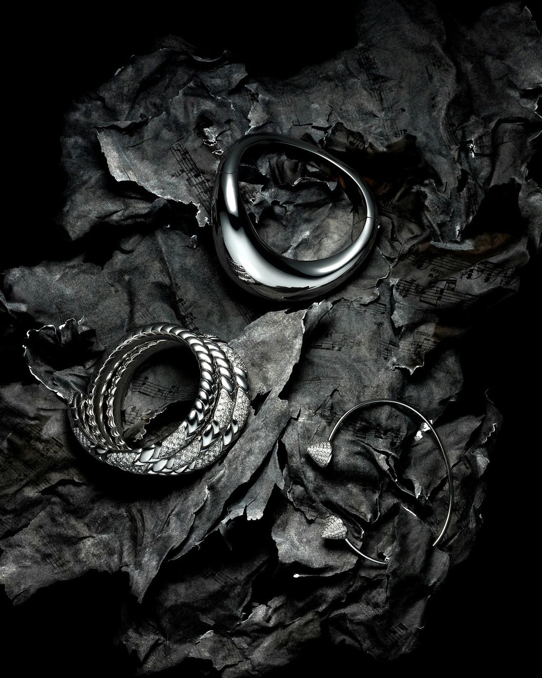 Silver rings and ear cuff jewelry arranged on crumpled sheet music paper with a dark, high-contrast fashion-style lighting scheme.