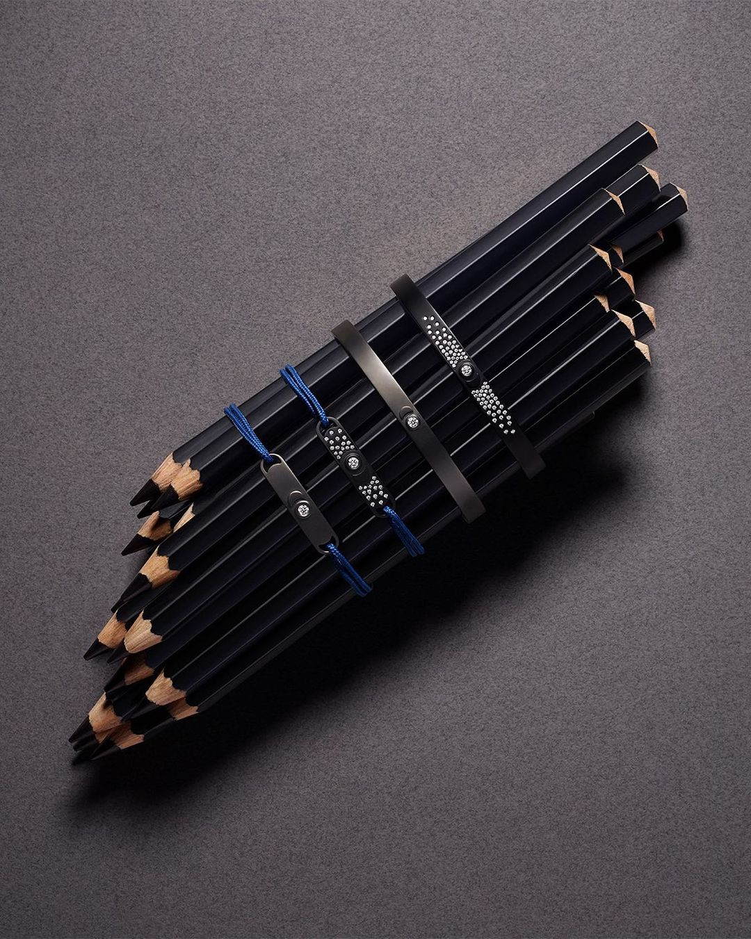A neatly arranged bundle of black pencils secured with decorative bands, displayed on a dark gray textured surface in a well-lit studio setting.