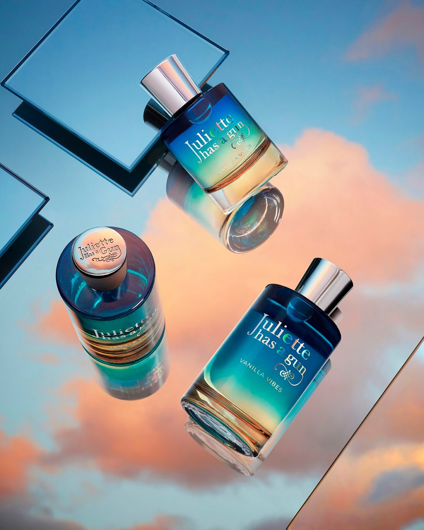 Three Juliette Has a Gun perfume bottles stand on a reflective surface with a blue, yellow, and pink sky reflected above.