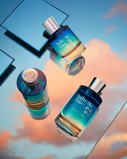 Three Juliette Has a Gun perfume bottles stand on a reflective surface with a blue, yellow, and pink sky reflected above.