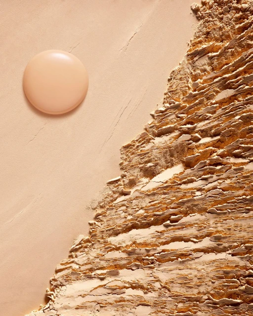 Beige-toned beauty product shot featuring a round container next to a textured surface resembling compacted powder or ground material.