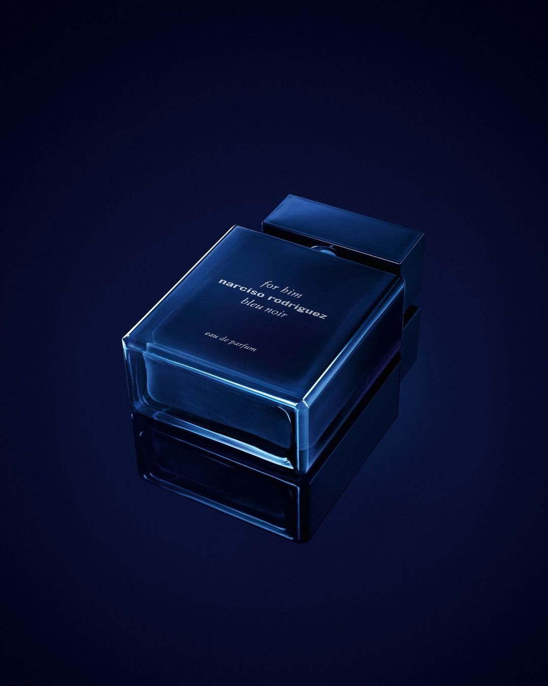 A rectangular dark blue perfume bottle resting on a dark reflective surface under moody blue lighting.